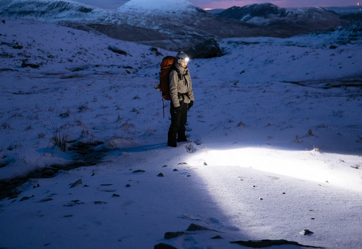 picture of person standing on a snow in mountains with a backpack and a flashlight 
