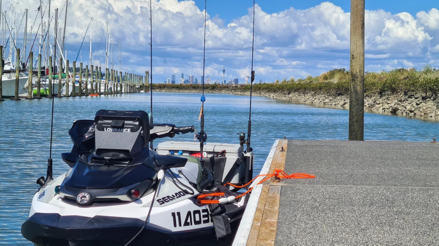 A Sea-Doo personal watercraft with fishing rods attached, docked at a marina with clear skies and city skyline in the background.